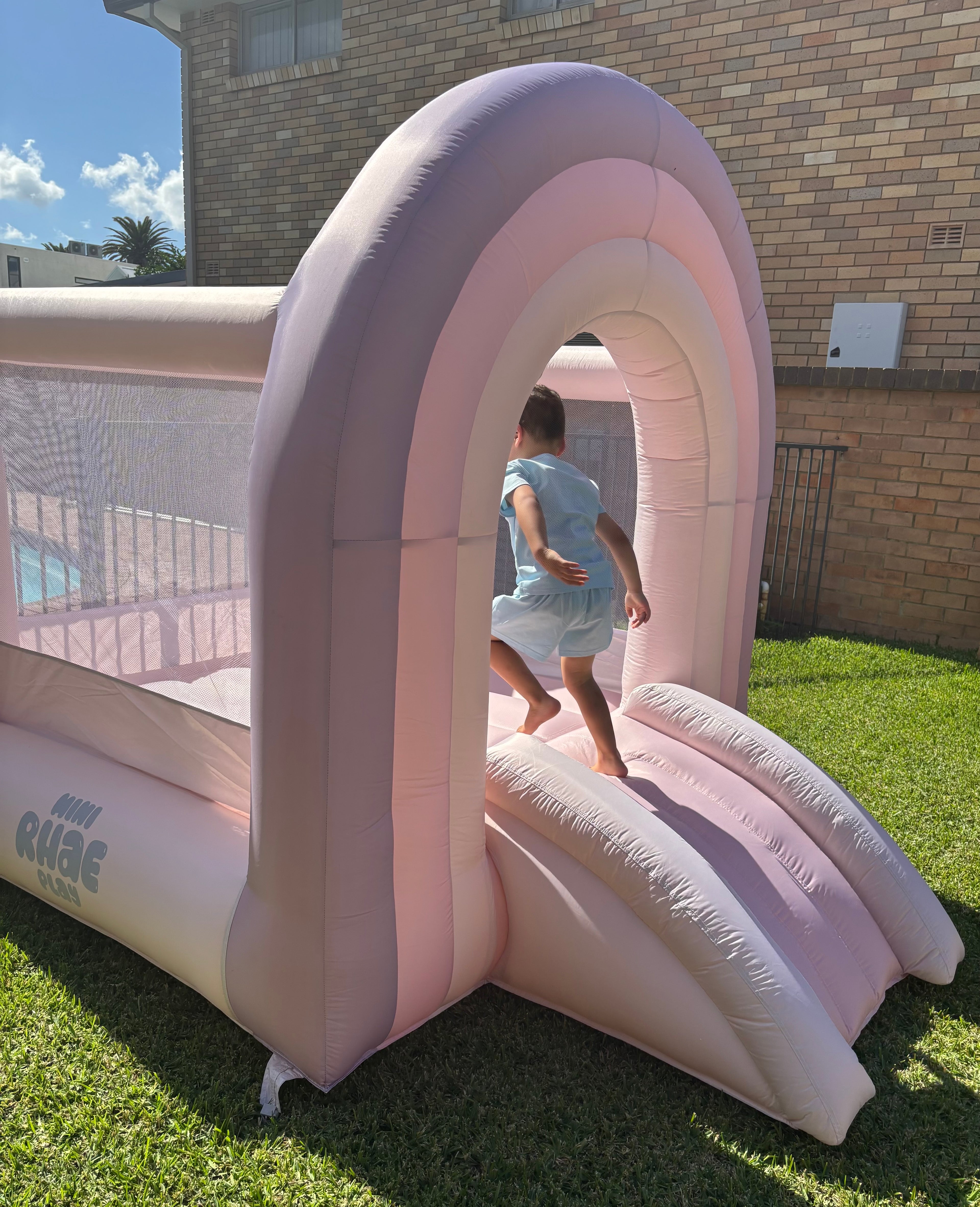 A child playing on a pink inflatable bouncer, which is set up on a grassy area outdoors.