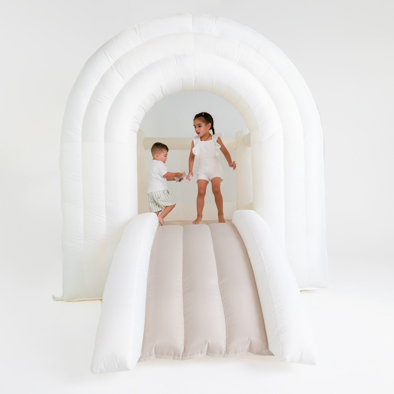 An inflatable bounce house with a rainbow arch design in white, featuring two children playing on it.